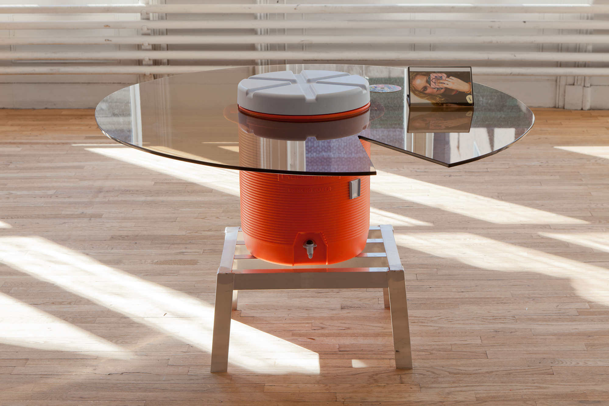 An orange water dispenser sits on a low metal stool in a gallery space. Encircling the top portion of the dispenser is a round glass table, with a wedge-shaped cut-out removed from its right side. A photo of a woman sits on top of the glass surface.