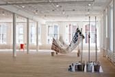 An installation view of a large room. To the left, an orange water dispenser is positioned between three white columns. In the center of the room is an abstract sculptural work. In the lower right foreground are eleven metal poles mounted in metal buckets that are filled with multicolored objects.