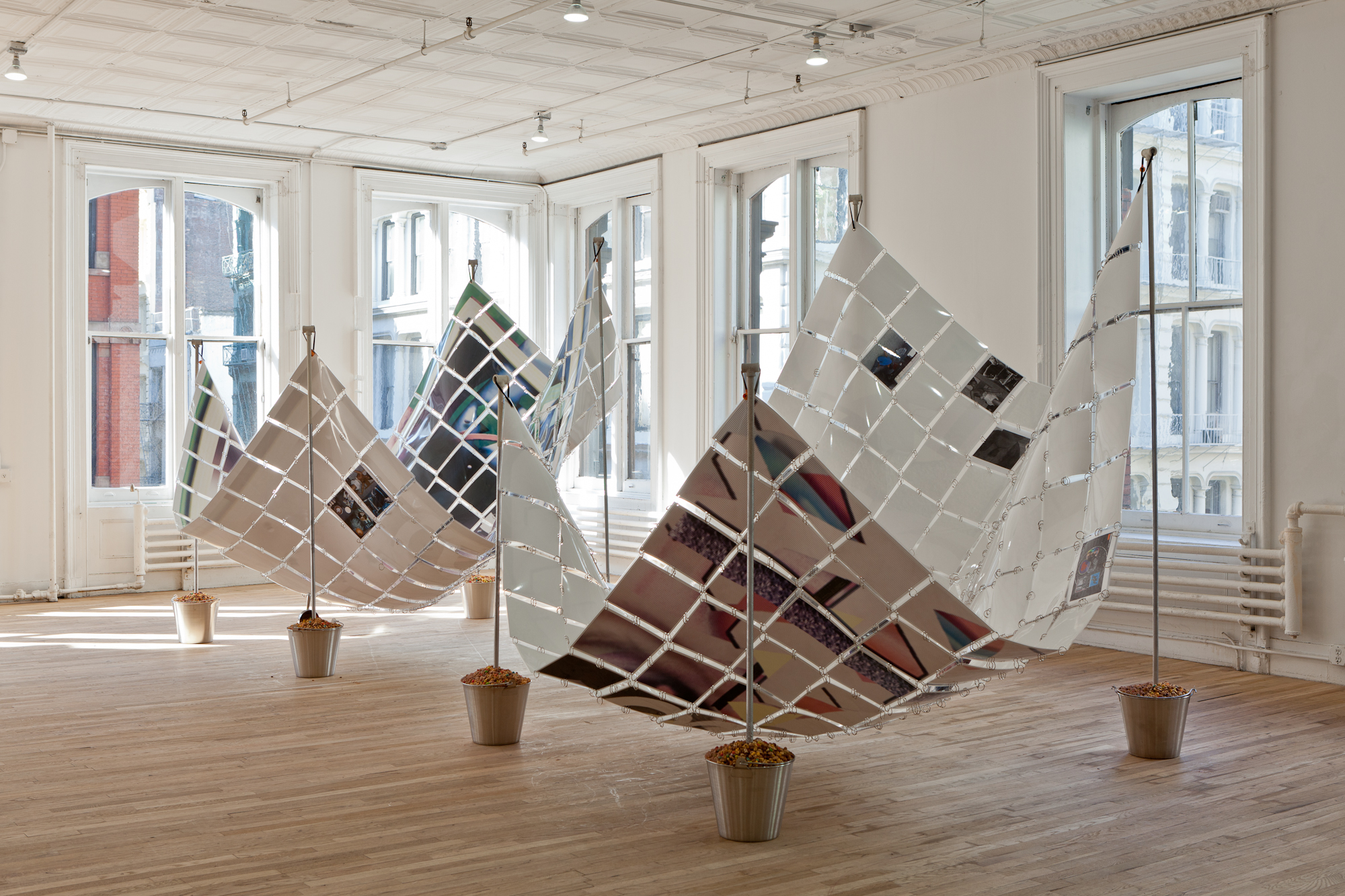 Two sculptures installed in a gallery space featuring rectangular sheets of laminated, paper rectangles held together by metal rings. The sheets are suspended at their four corners by metal poles mounted in silver buckets.