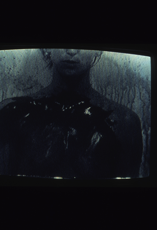 Dark image of the upper torso of a woman with liquid dripping in the foreground of the image.