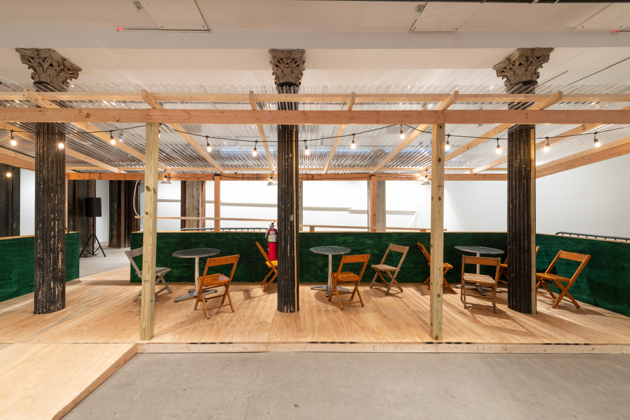 An installation view of a green and tan wooden dining corral with a ramp, metal tables and wooden chairs.