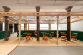 An installation view of a green and tan wooden dining corral with a ramp, metal tables and wooden chairs.