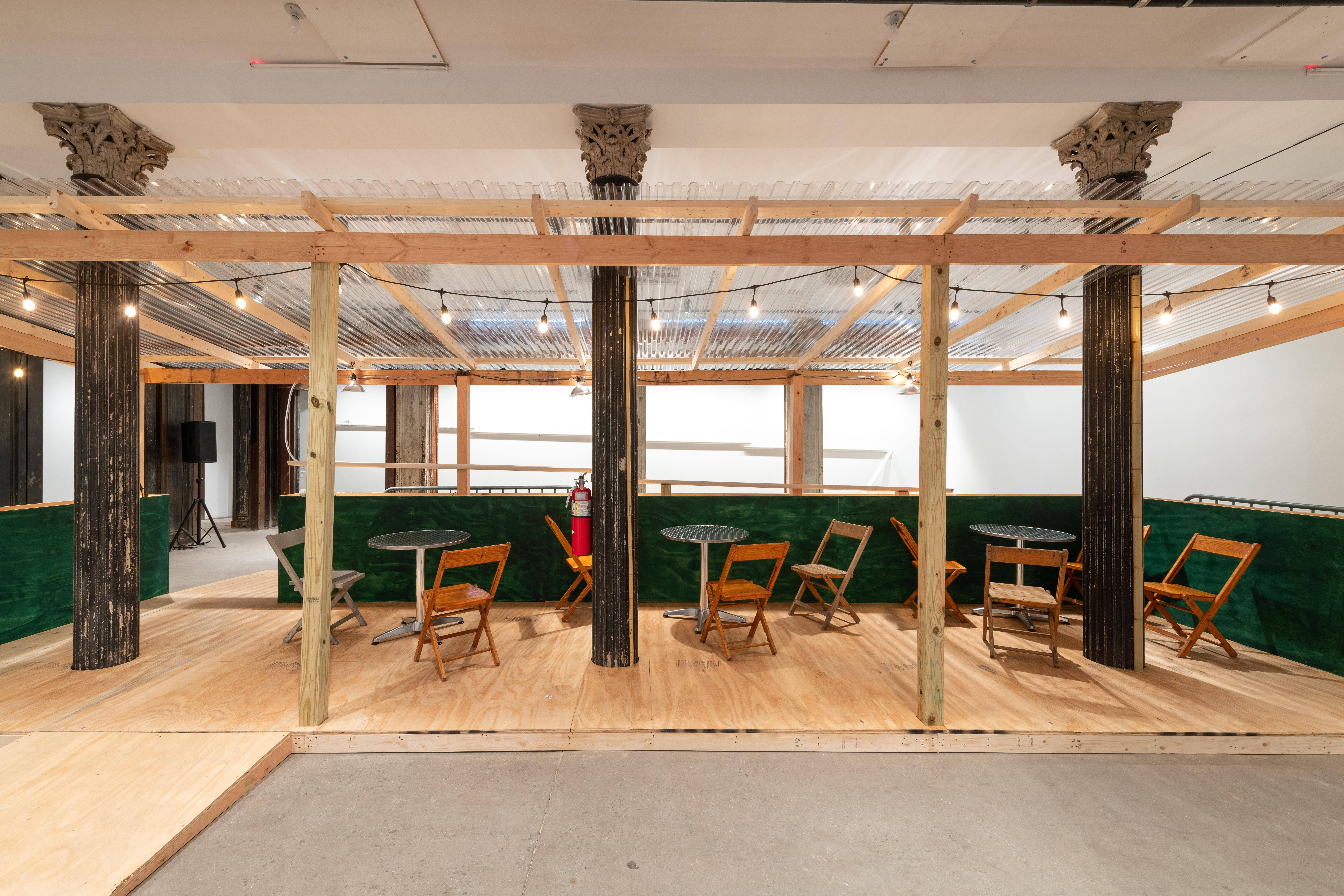 An installation view of a green and tan wooden dining corral with a ramp, metal tables and wooden chairs.