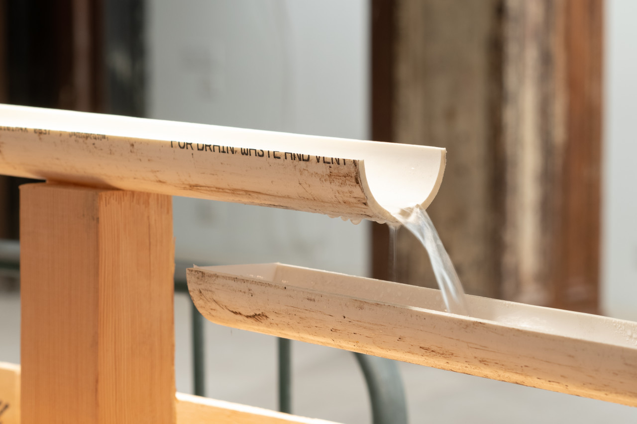 A sharp image of two halves of a pvc pipe placed in front of an out of focus background. Water flows across the pipes; behind the pipes is a steel column.