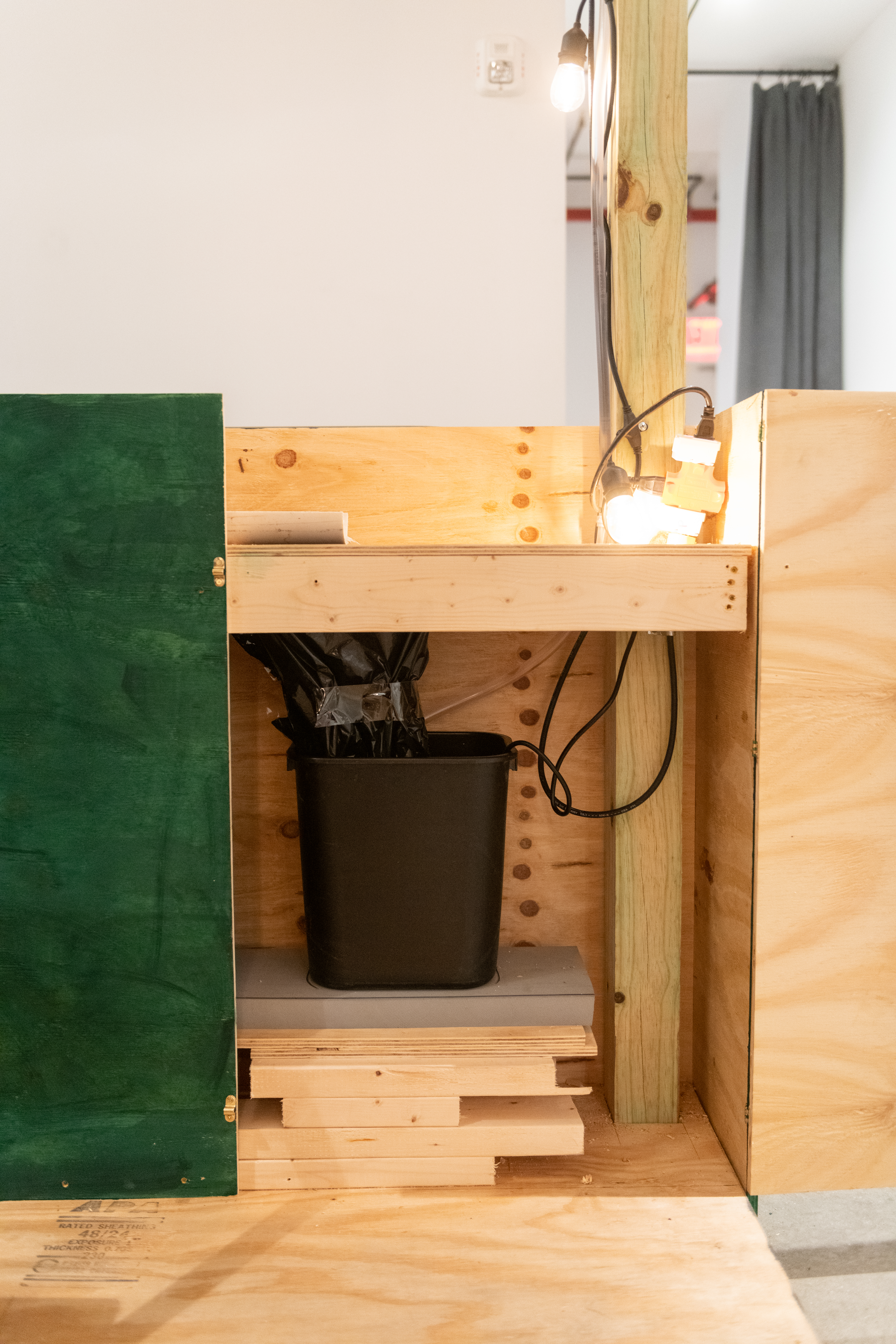 An installation view of the corner of a green and tan wooden dining corral. In the center a black plastic bin with black tubes and crumpled black fabric like material is visible.