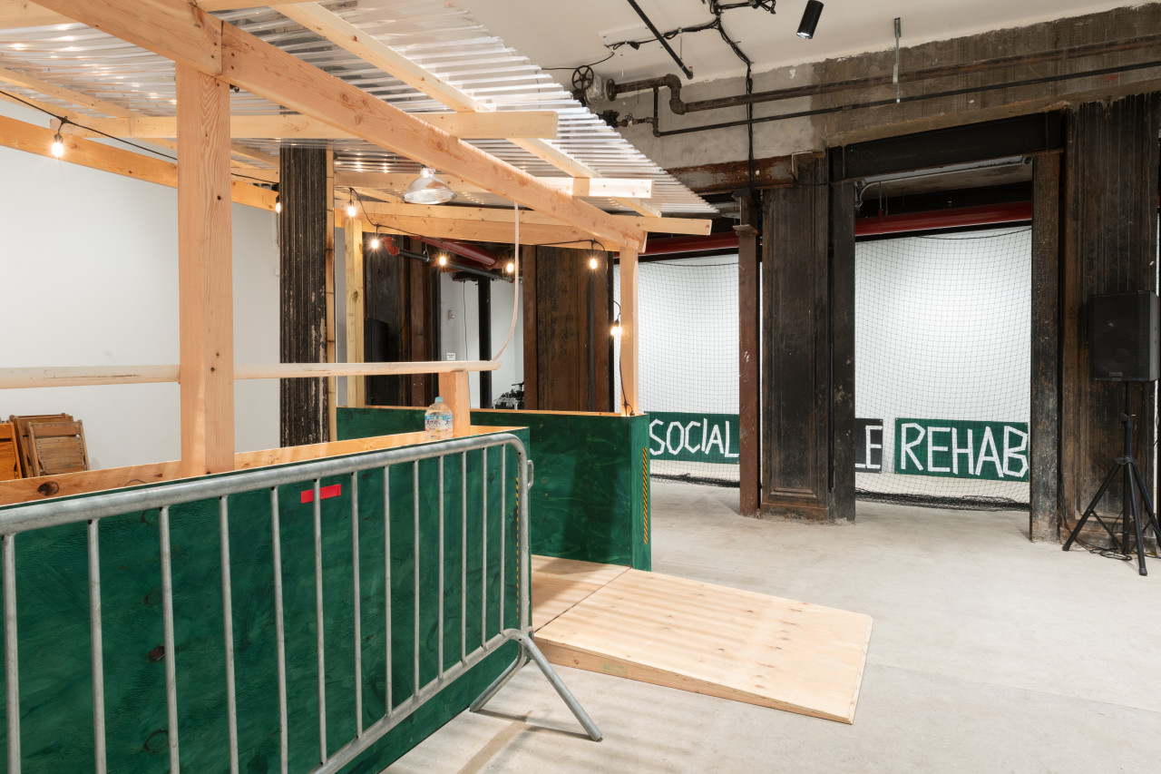 Installation view of a gallery space with a green and tan wooden dining corral with a metal barricade. In the background are four grey concrete columns. Between the gaps of the columns a net and two signs which read "Social" and "Rehab" are visible.