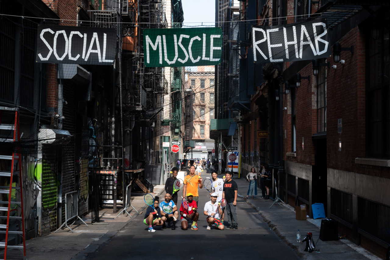 A group of eight people holding tennis rackets stand and crouch in an alleyway. On signs suspended above them, the words "Social Muscle Rehab" are painted in white capitalized text over black and green backgrounds.