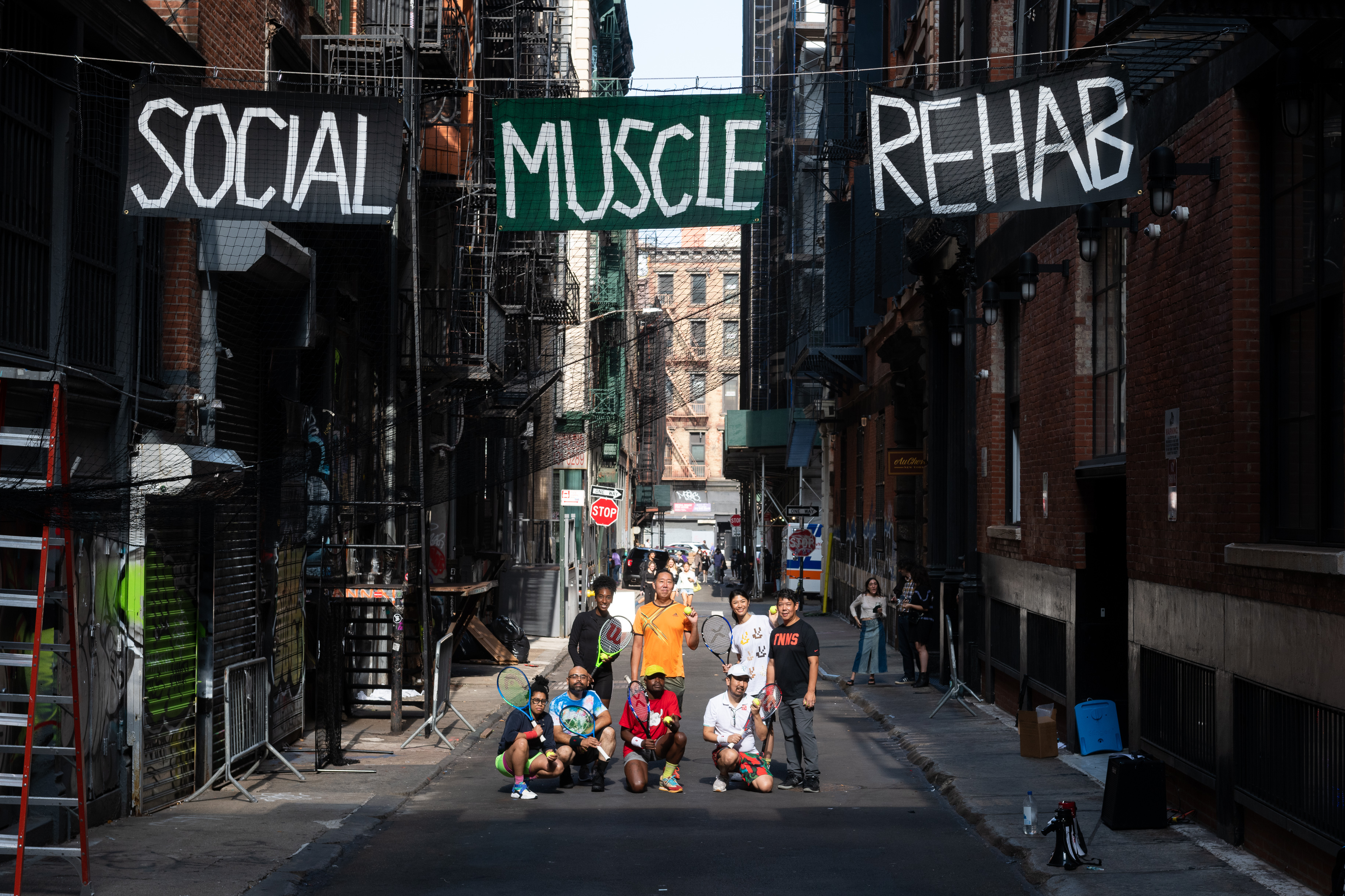 A group of eight people holding tennis rackets stand and crouch in an alleyway. On signs suspended above them, the words 