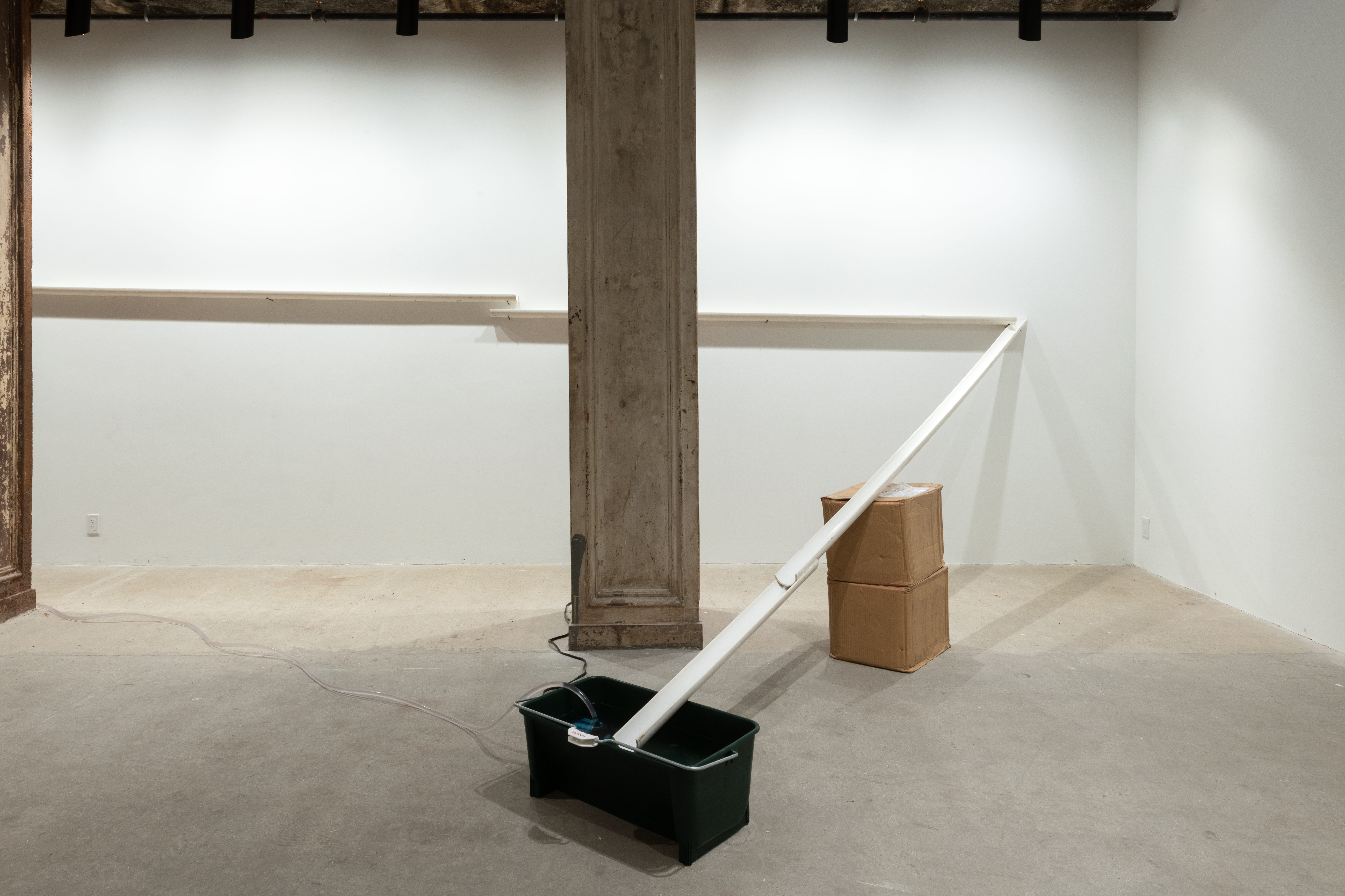 Installation view of white PVC pipe that is cut in half lengthwise. The pipe runs along a wall in the background and makes a sharp turn to drain into a black plastic bin on the floor.