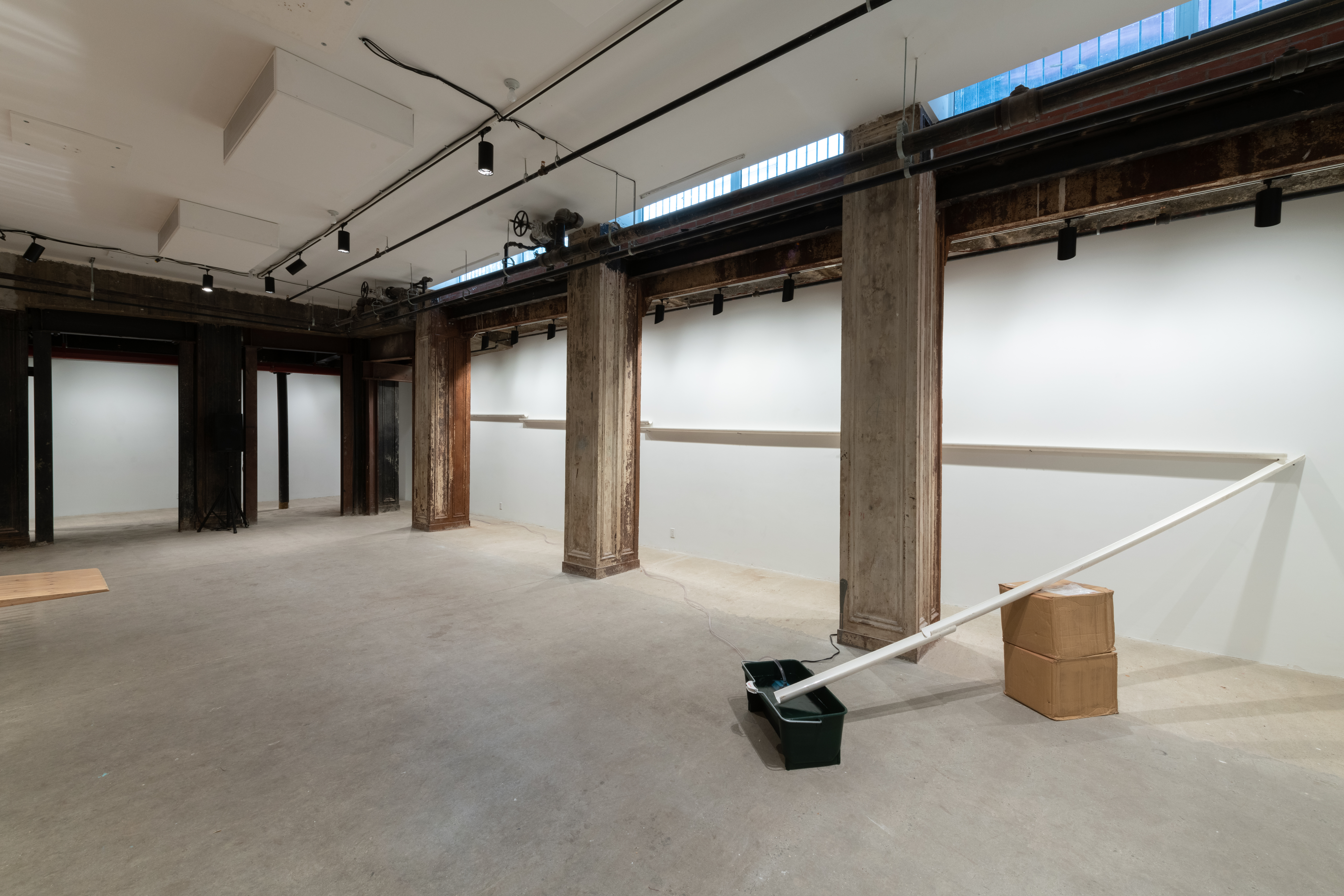 Installation view of white PVC pipe that is cut in half lengthwise. The pipe runs along a wall on the right and makes a sharp turn to drain into a black plastic bin on the floor.