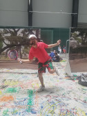 A person in a red shirt, white cap, and multicolored pants smiles and poses with a tennis racquet, standing atop a large piece of paper with colorful drawings on it. In the background are people sitting on the ground and printed images of trees.