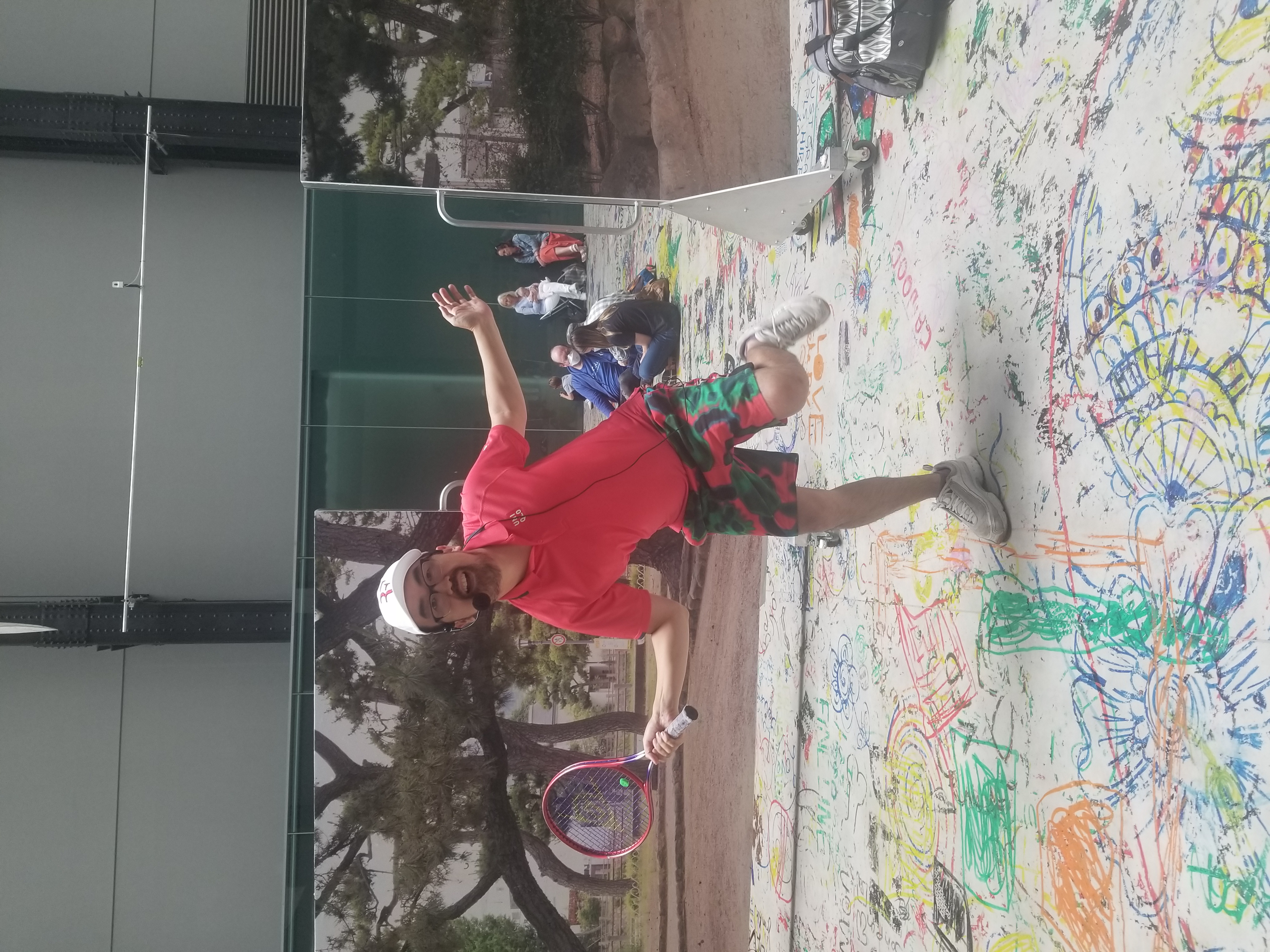 A person in a red shirt, white cap, and multicolored pants smiles and poses with a tennis racquet, standing atop a large piece of paper with colorful drawings on it. In the background are people sitting on the ground and printed images of trees.