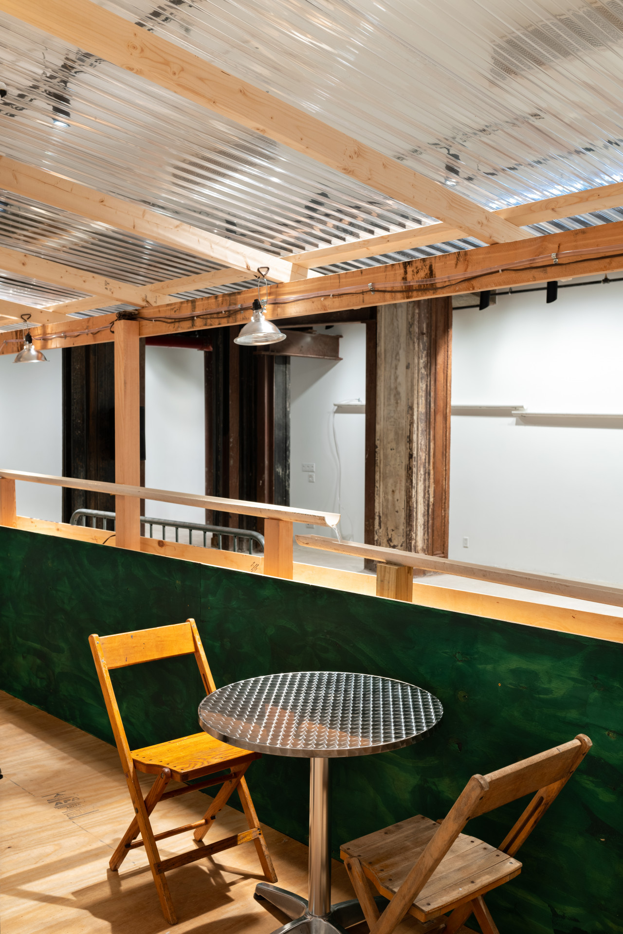 An installation view of a green and tan wooden dining corral with a metal table and two wooden chairs. Halved PVC pipes are visible in the background.