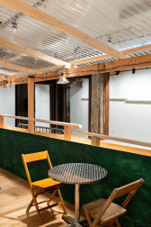 An installation view of a green and tan wooden dining corral with a metal table and two wooden chairs. Halved PVC pipes are visible in the background.