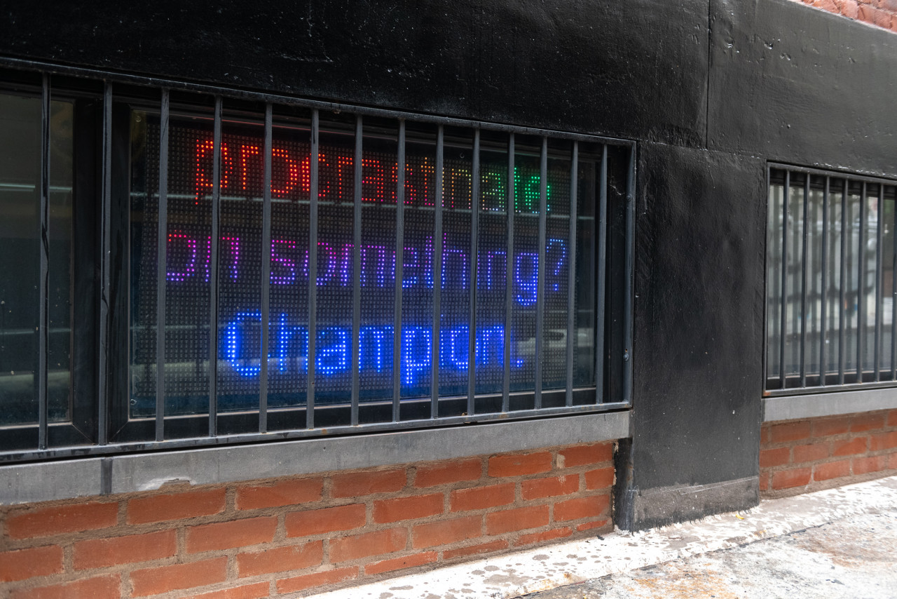 An LED screen is visible behind a set of black bars at the level of a sidewalk. The screen reads in multicolored text "procrastinate on something? Champion."