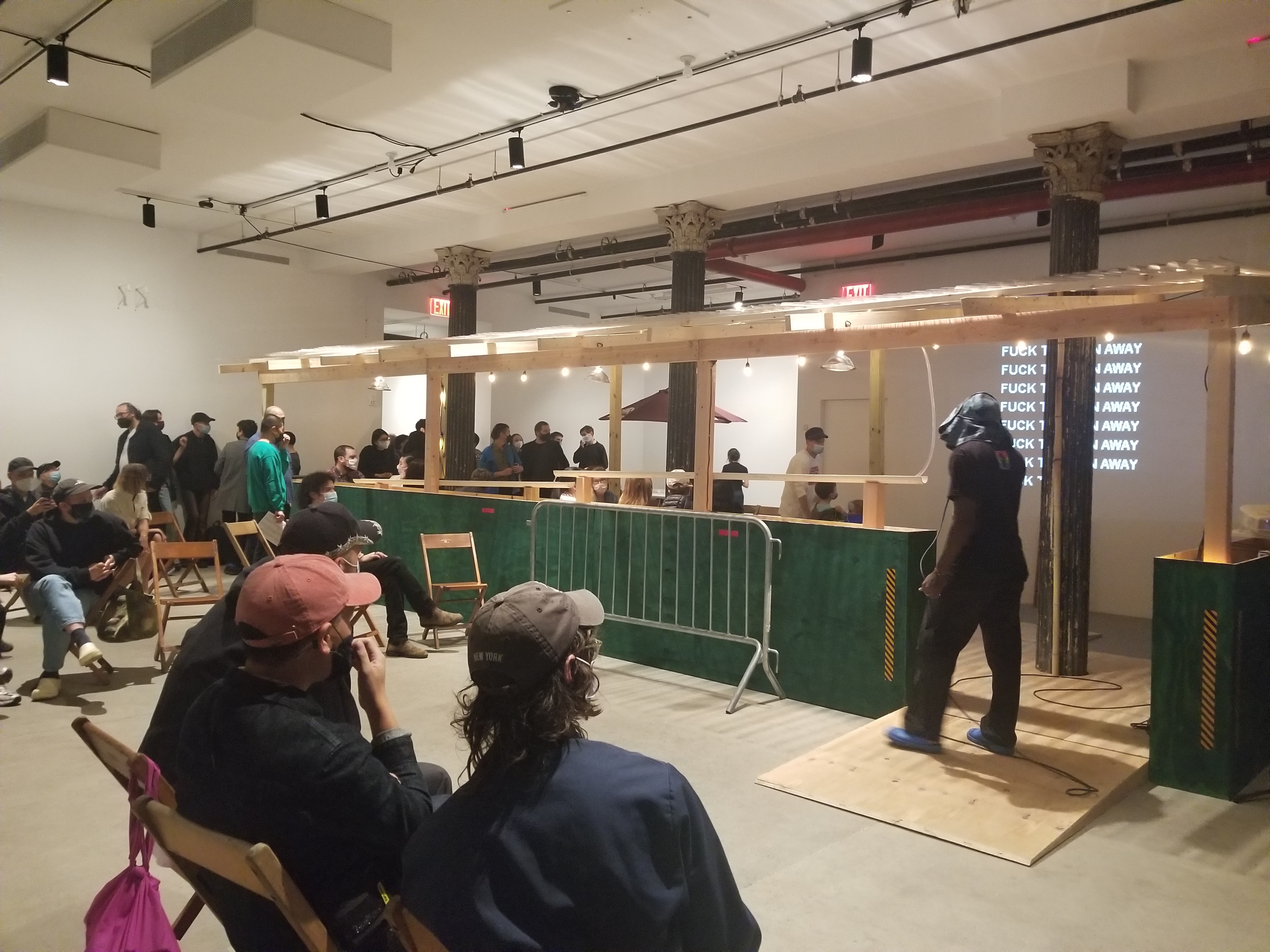 A green and tan wooden dining corral with a ramp and a metal barricade. People sit and stand in and around the corral.