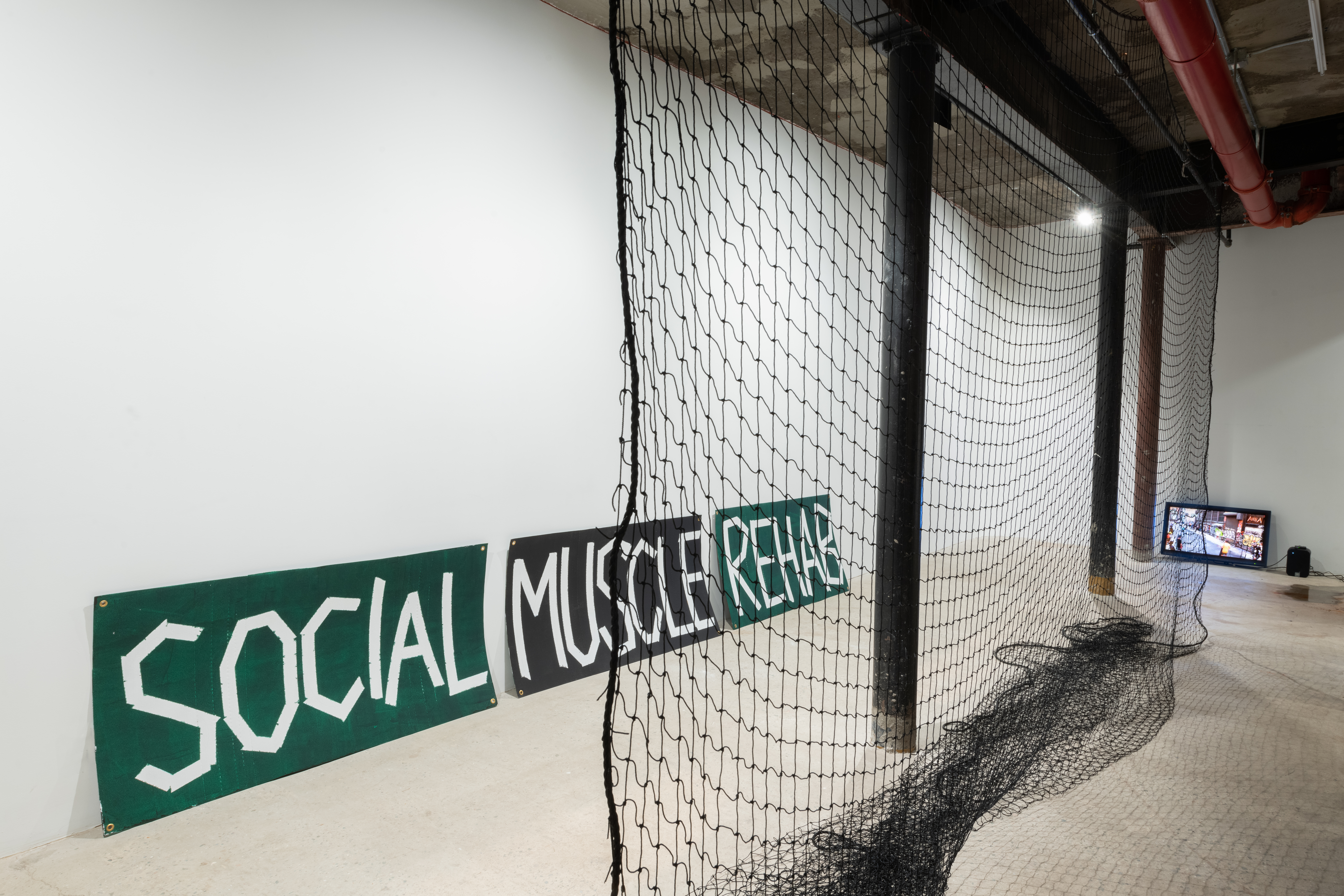 View of a gallery space. A black net hangs from the ceiling, extending from the center foreground into the background where a small television screen is visible. On the left, three signs are propped against the wall. In white text over green and black backgrounds the signs read 