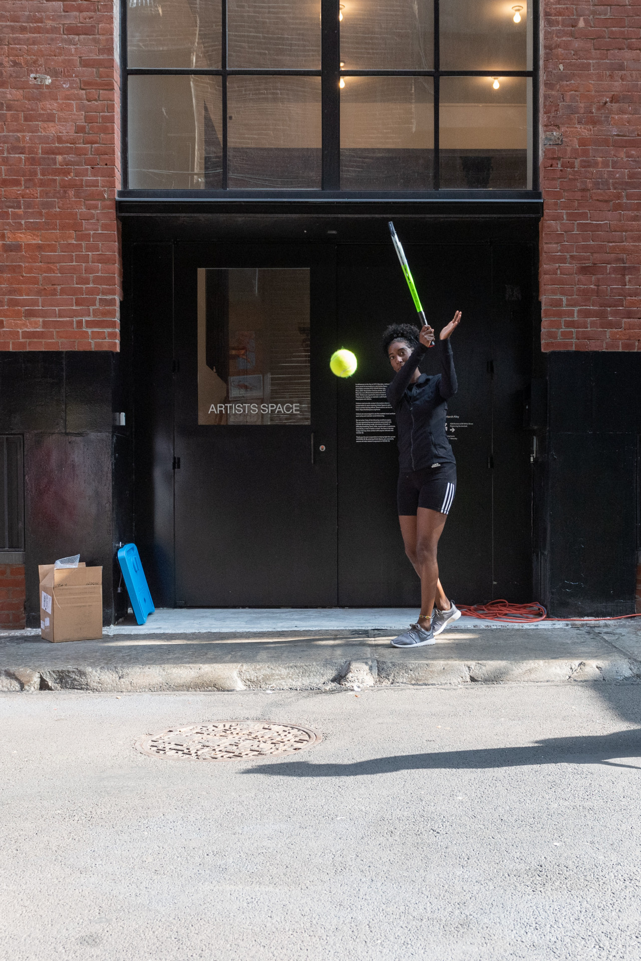 A person in black athletic wear hits a tennis ball towards the camera in an alleyway. Behind them, the words "Artists Space" appear in white text on the windowpane of a black door.