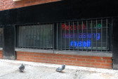 An LED screen can be seen behind a set of black bars at the level of a sidewalk. The screen reads in multicolored text, 