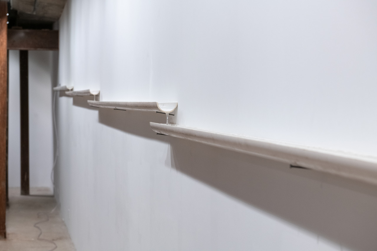Installation view of white PVC pipes that are cut in half lengthwise. The pipes rest on nails against the wall and water trickles inside.