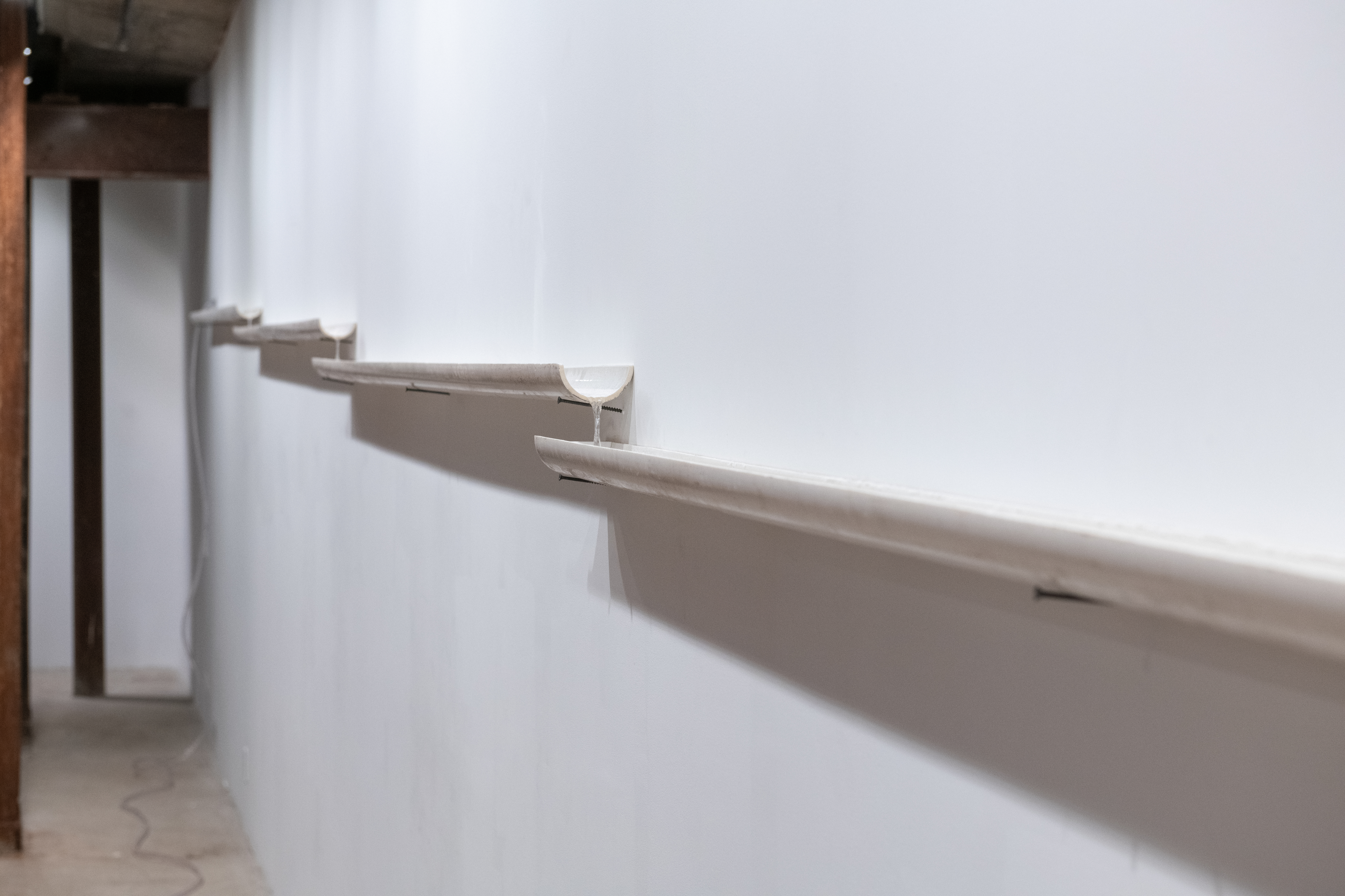 Installation view of white PVC pipes that are cut in half lengthwise. The pipes rest on nails against the wall and water trickles inside.