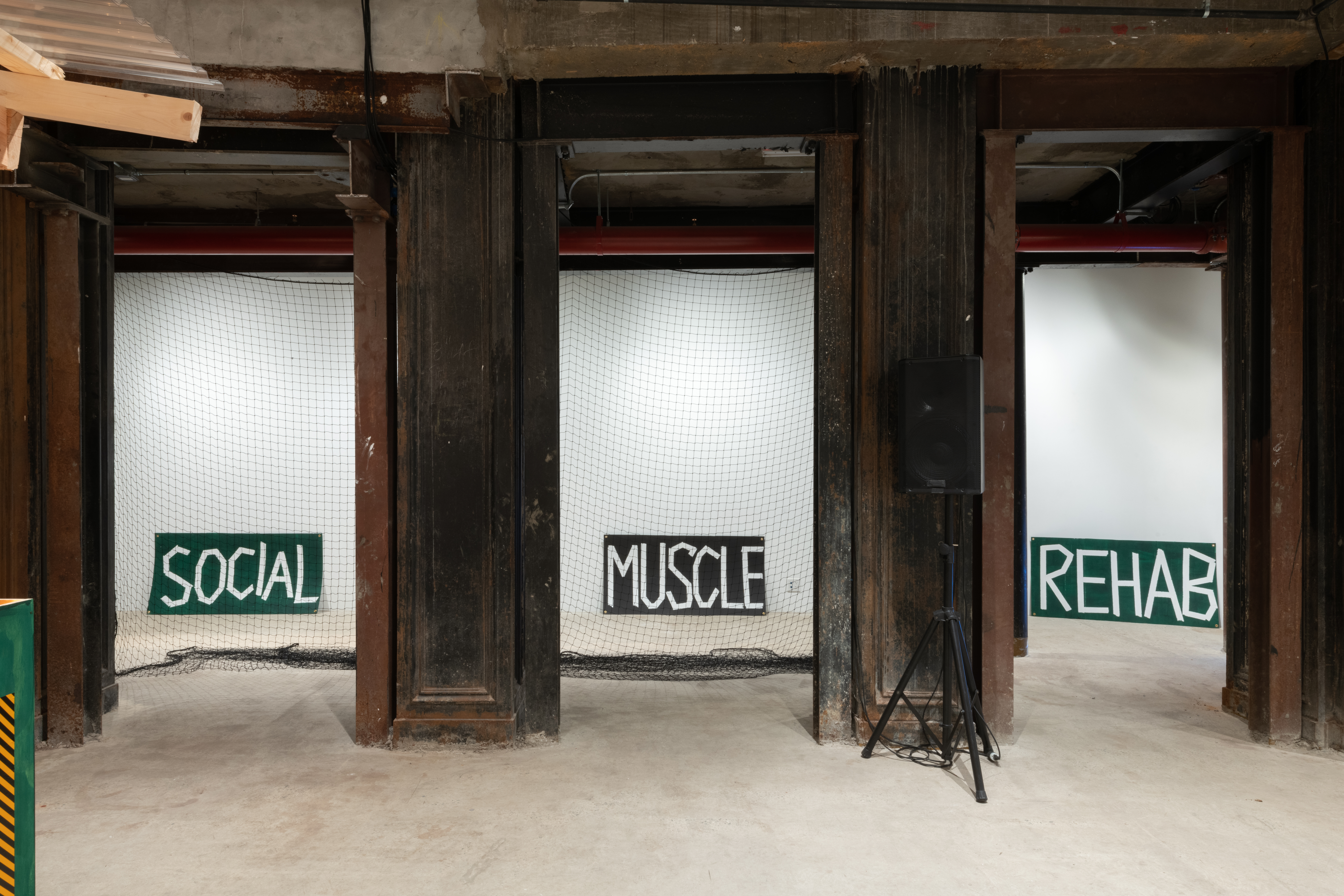 View of a gallery space with four grey concrete columns. In the gaps between the columns, a net and three signs are visible. The signs read, 