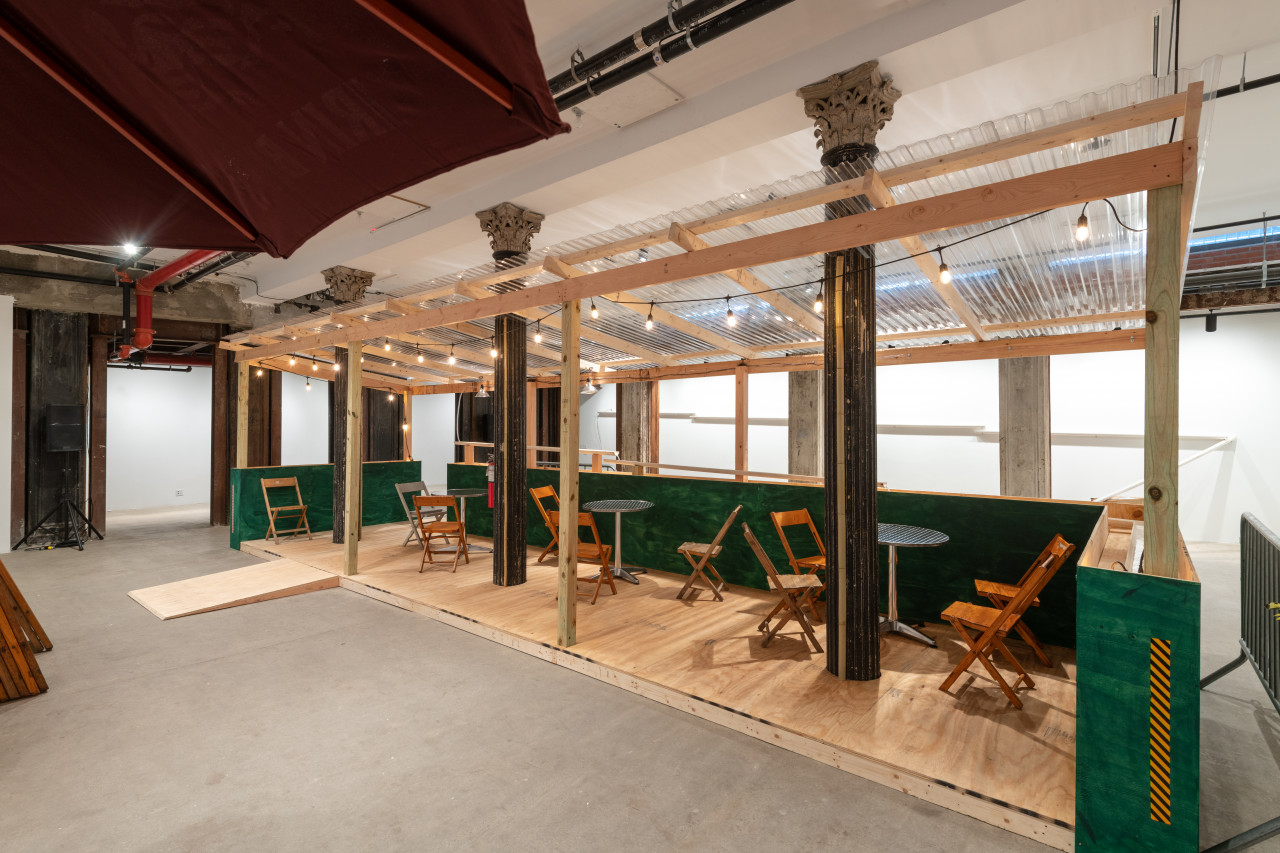 An installation view of a green and tan wooden dining corral with a ramp, metal tables, and wooden chairs.
