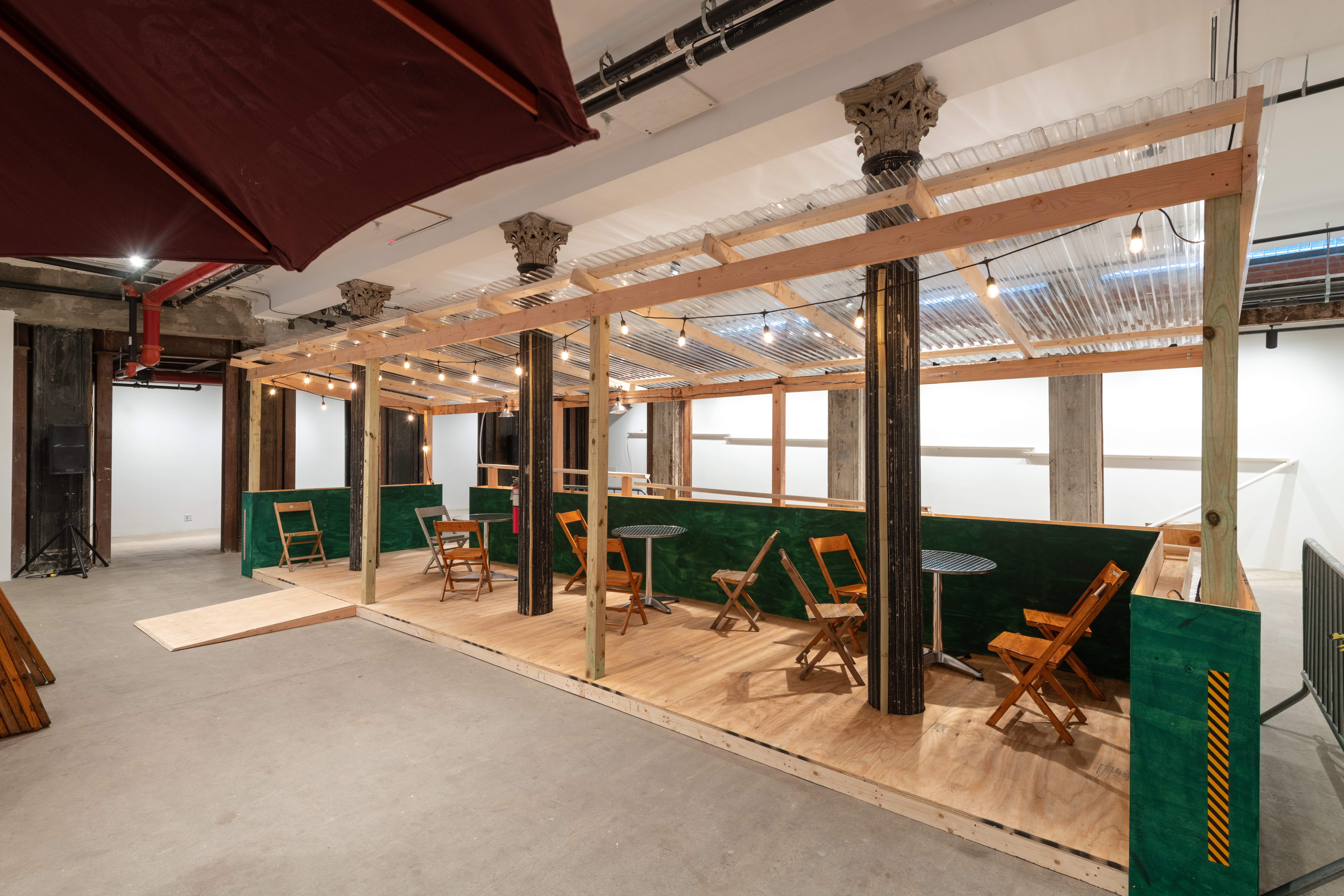 An installation view of a green and tan wooden dining corral with a ramp, metal tables, and wooden chairs.