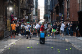 Six people with tennis rackets in an alleyway jump and swing to hit tennis balls towards the camera which is behind a black net. A crowd behind the tennis players watches attentively.