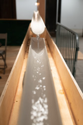 Image of a PVC pipe that has been cut in half lengthwise, nestled in the wall of a wooden dining corral. Water flows through the pipe towards the camera. On the right, a metal barricade can be seen.