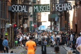 Tennis players stand in an alleyway, ready to hit balls from an automatic ballfeeder. A crowd watches intently in the background. Suspended above them the words 