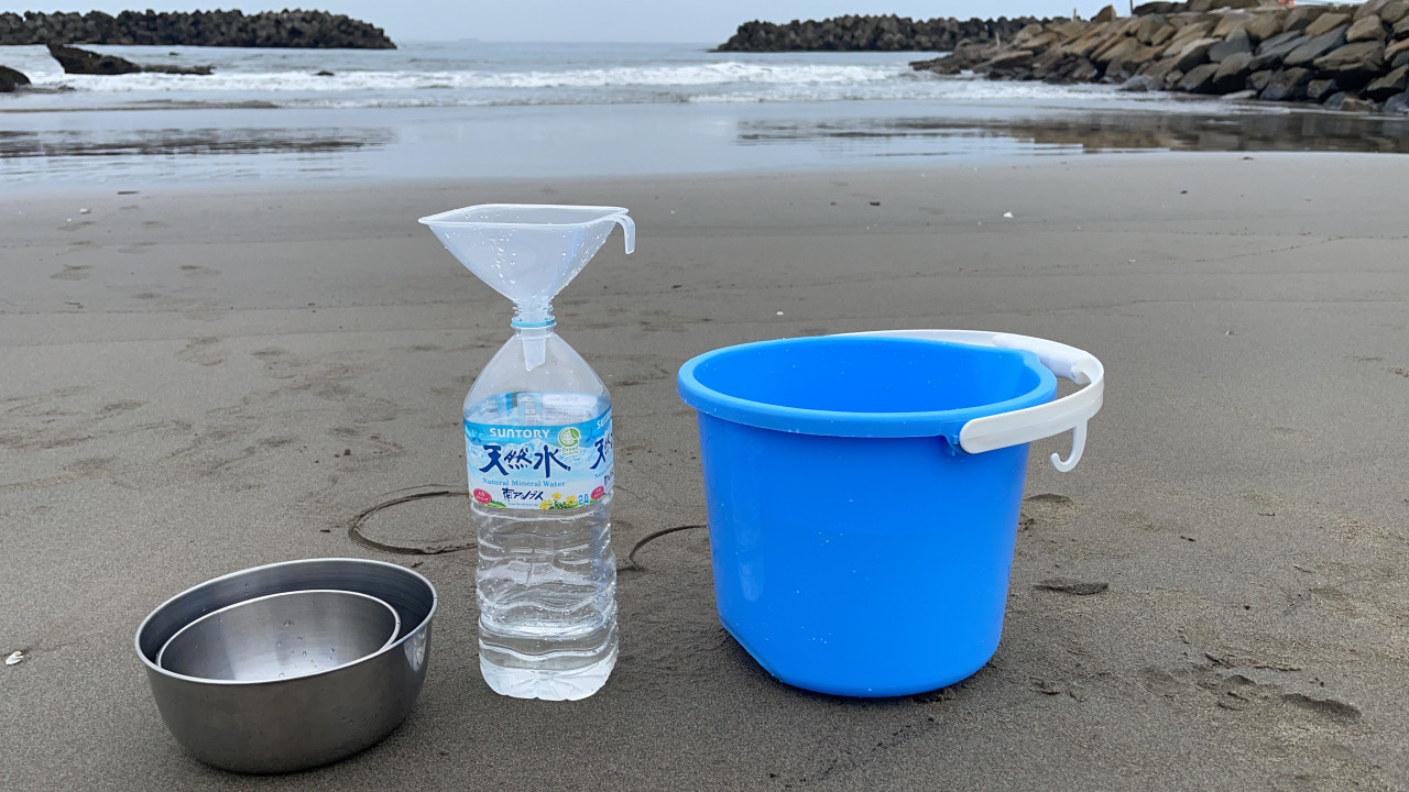 Two metal bowls, a water bottle with a funnel in its spout, and a blue plastic bucket stand on a sandy ocean beach