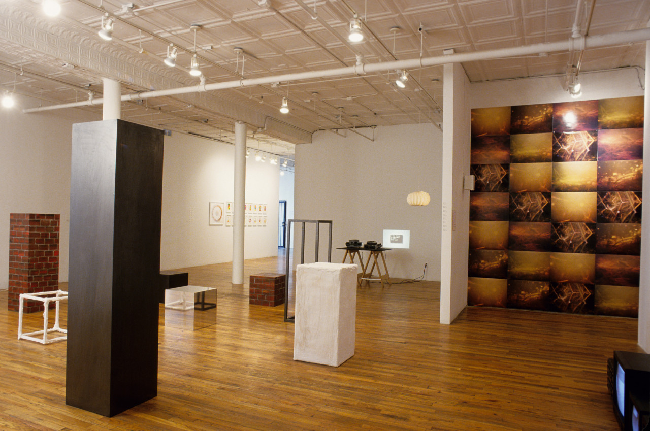 A gallery room filled with various sculptures and objects. There are several cube and rectangular sculptures in various sizes, some solidly painted with black, white and silver, and some painted with a brick pattern. There is a table with two projectors projecting a small image on the wall. On the far wall, there are a series of framed drawings. On the righthand side of the room, there is a half-wall behind which a piece made of 7x4 rectangular images hangs on a wall.