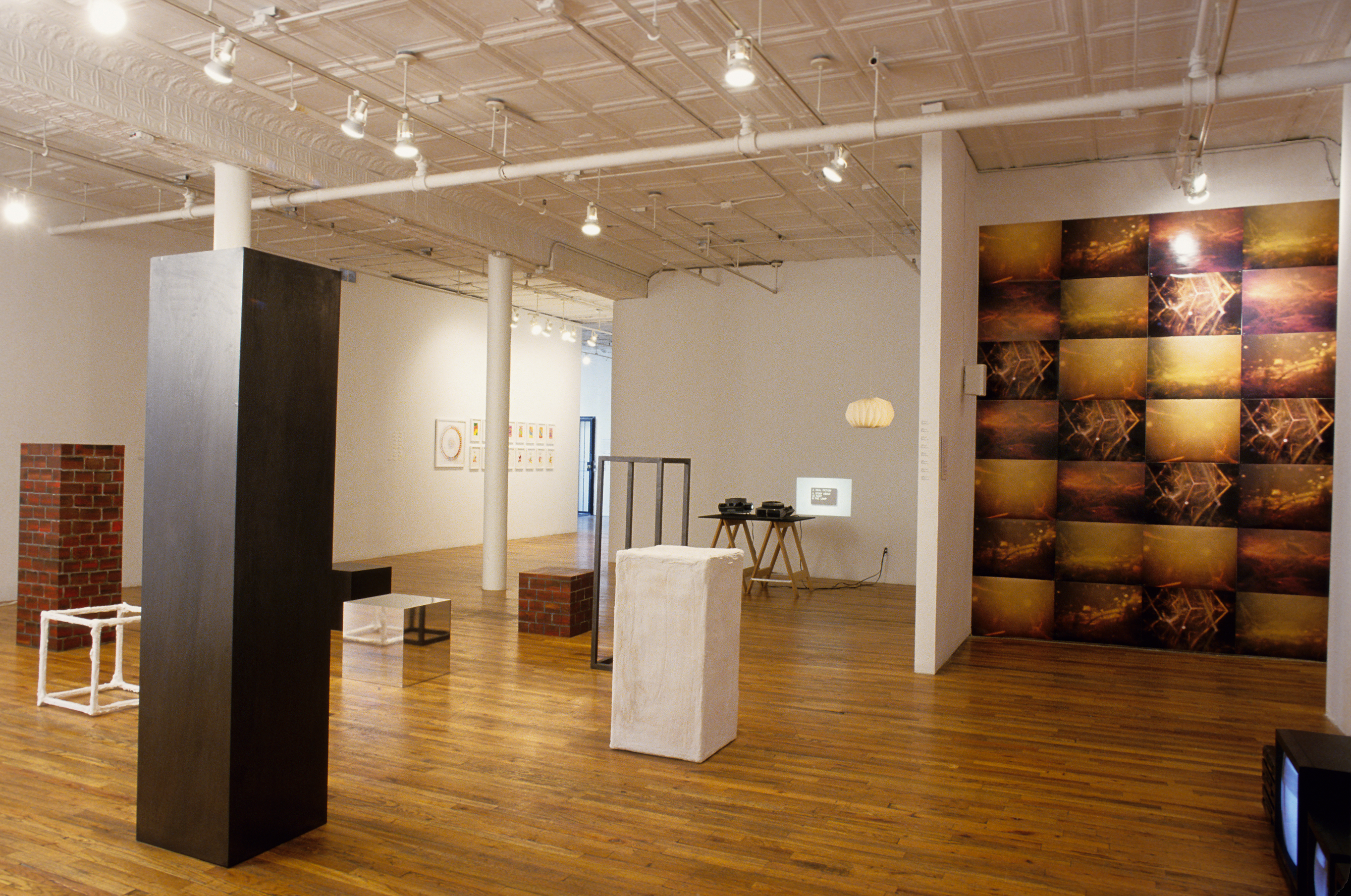 A gallery room filled with various sculptures and objects. There are several cube and rectangular sculptures in various sizes, some solidly painted with black, white and silver, and some painted with a brick pattern. There is a table with two projectors projecting a small image on the wall. On the far wall, there are a series of framed drawings. On the righthand side of the room, there is a half-wall behind which a piece made of 7x4 rectangular images hangs on a wall.