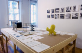 A wide wooden worktable sits in a gallery space with various models, books, and papers displayed on its surface. One of the models located closest to the viewer is yellow and has an amorphous shape. A series of black and white photographs of buildings hang on the wall to the right of the table.