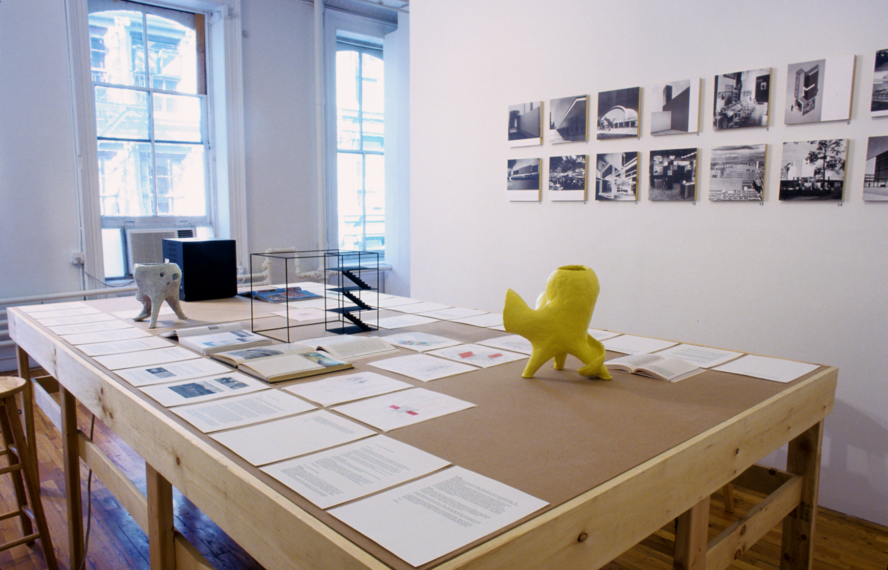 A wide wooden worktable sits in a gallery space with various models, books, and papers displayed on its surface. One of the models located closest to the viewer is yellow and has an amorphous shape. A series of black and white photographs of buildings hang on the wall to the right of the table.