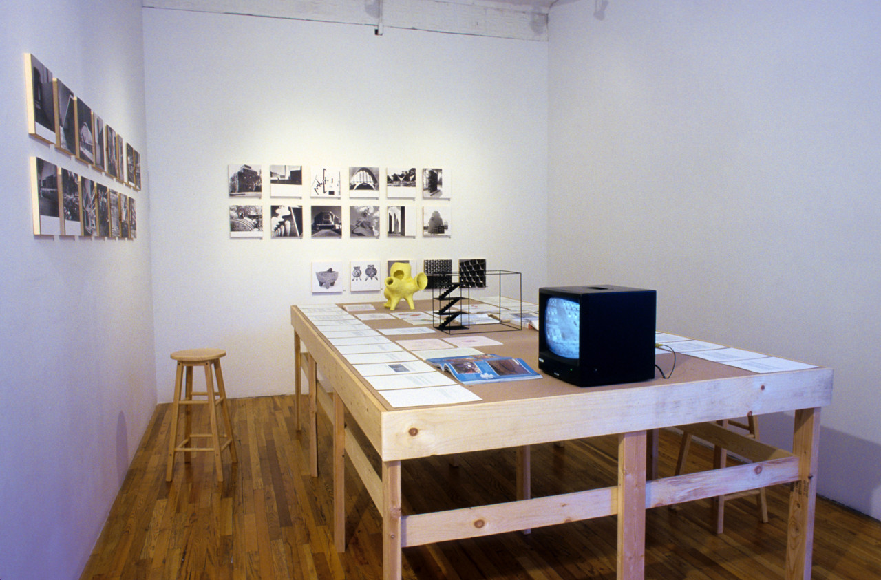 A wide wooden worktable is installed in a gallery space. Various papers, books, models, and a small television set are displayed on the table. A series of black and white photographs are installed on the two gallery walls that surround the table.