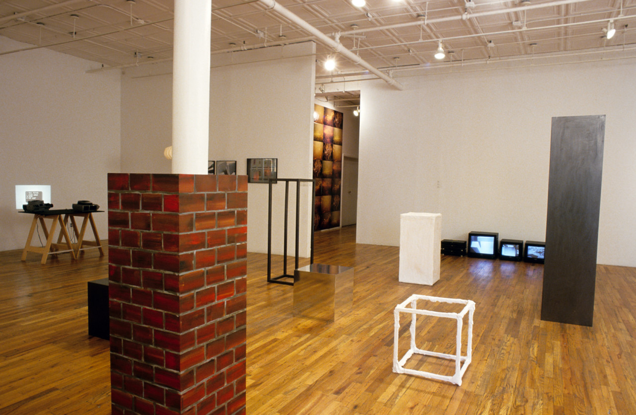 A gallery room filled with various sculptures and objects. There are four plinths of varying heights with black, white, silver, and mirrored surfaces. There are three televisions sitting on the floor against a white wall. On the other side, there are two projectors sitting on a table projecting small images on white walls. In the foreground, a rectangular cube painted like bricks is wrapped around a column.