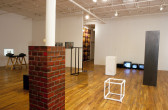 A gallery room filled with various sculptures and objects. There are four plinths of varying heights with black, white, silver, and mirrored surfaces. There are three televisions sitting on the floor against a white wall. On the other side, there are two projectors sitting on a table projecting small images on white walls. In the foreground, a rectangular cube painted like bricks is wrapped around a column.