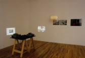 Two slide projectors sit on a table, projecting small images onto perpendicular walls. On one wall, there is a paper lamp and three framed photographs: two black and white images of folded paper, and one color image.
