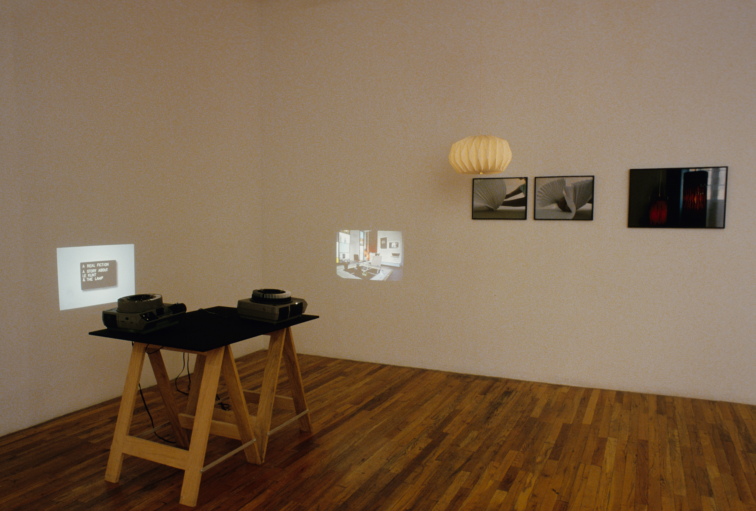 Two slide projectors sit on a table, projecting small images onto perpendicular walls. On one wall, there is a paper lamp and three framed photographs: two black and white images of folded paper, and one color image.