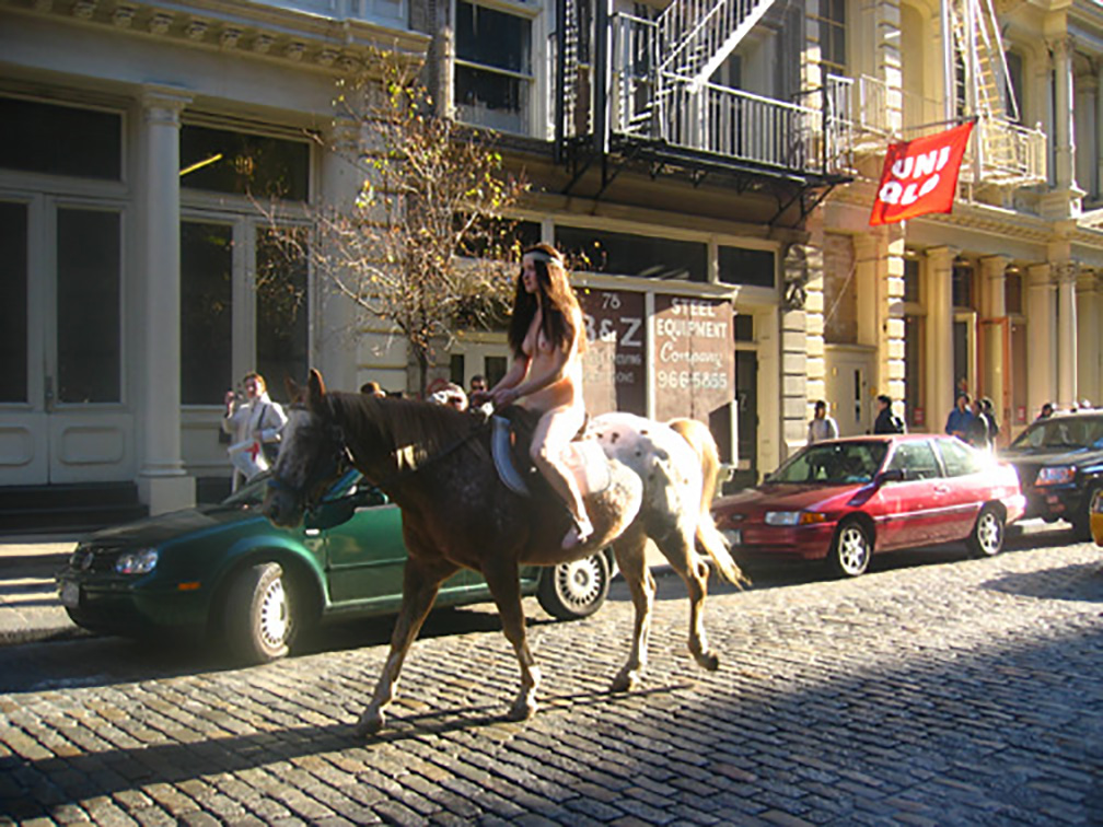 A naked woman with long, brown hair rides on a saddled brown and white horse down a cobbled urban street. One green and one red car are parked on the side of the street directly behind her, and various passerby look up towards her as she passes by. A red flag with white text reading, "UNIQLO" hangs from a rightmost building behind the horse.