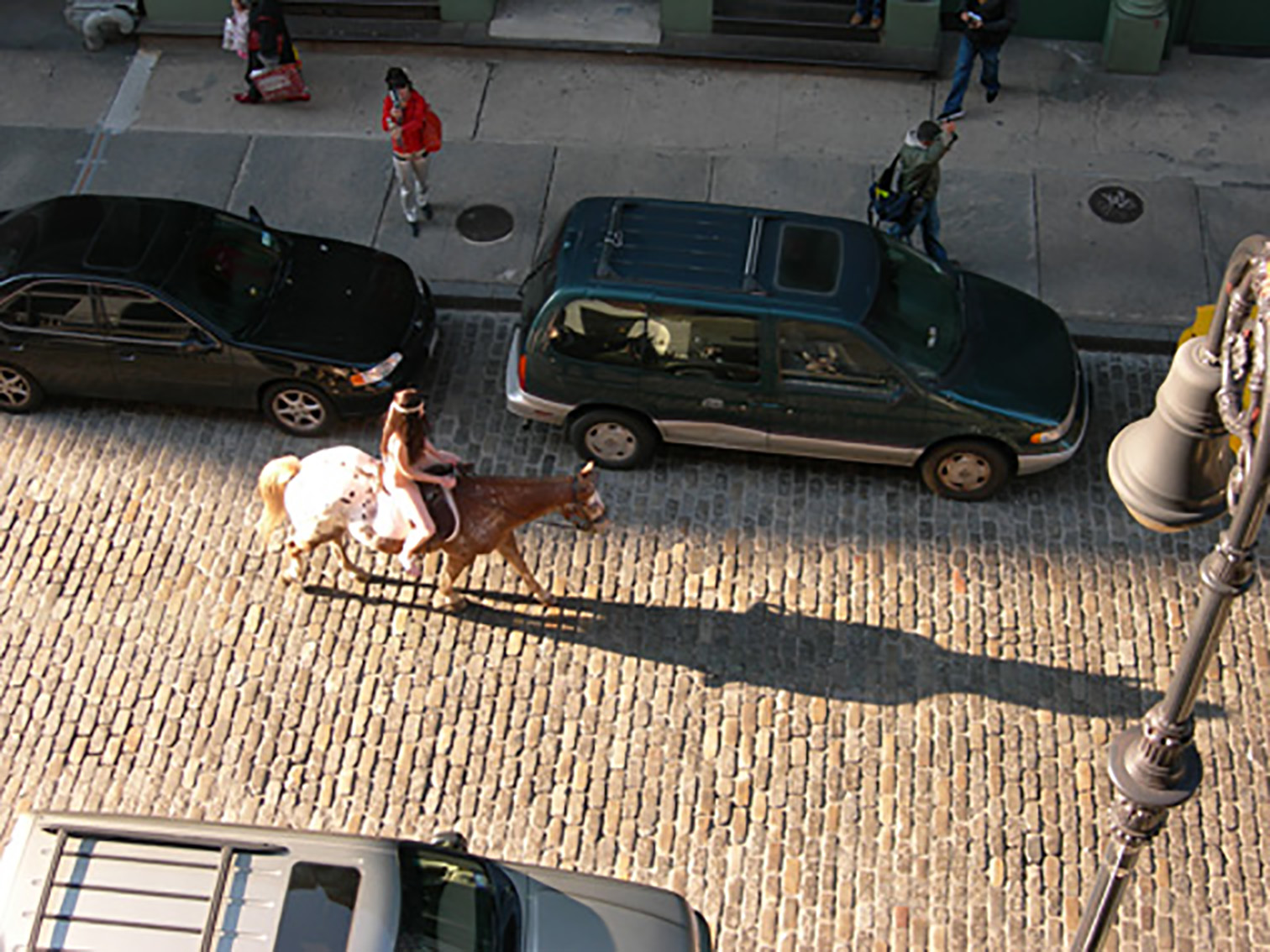 An aerial view of a naked woman with long, brown hair riding on a saddled brown and white horse down a cobbled urban street. There are parked cars on either side of her, a single lampost, and a few pedestrians looking on. She casts a long shadow onto the cobbled street.