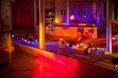 A installation wide shot of a room with five silver columns. In the foreground, there is a rectangular pool filled with water with two spouts spraying water in fountains. A second pool built from wood is visible in the background. Between the two, there are various objects including a low-hanging disco ball, a vase with flowers, strands of string lights, and more. On the back wall, the word 