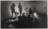 A black and white photograph showing a room filled with various standing lights and assorted objects sitting on the floor. Two people are standing, their outlines casting shadows on a white wall with the word 