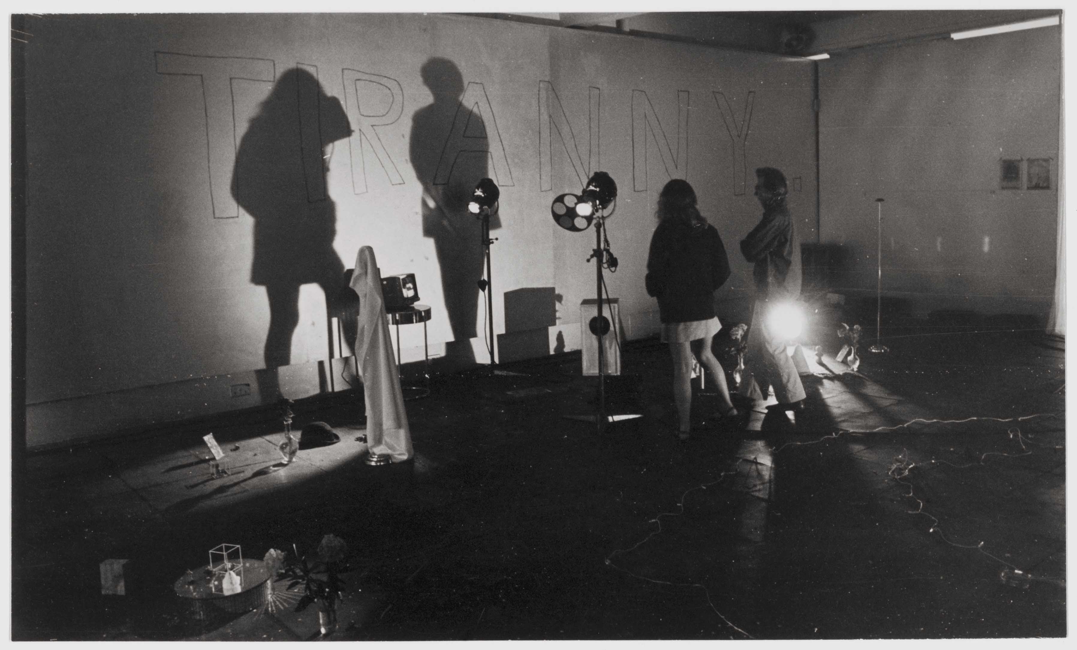 A black and white photograph showing a room filled with various standing lights and assorted objects sitting on the floor. Two people are standing, their outlines casting shadows on a white wall with the word 