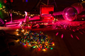 A pile of multicolored string lights sits on a wooden floor, surrounded by assorted objects including a disco ball, a candlestick, and two pitchers. There is a pink glow over the objects, with another strand of white lights weaving through the objects.