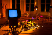 Objects are cluttered on the floor of a room lit with a warm, yellow hue. There is a low-hanging disco ball, two standing lights, and a small television showing white static mounted on a stand. There are two vases with plants and a string of white lights.