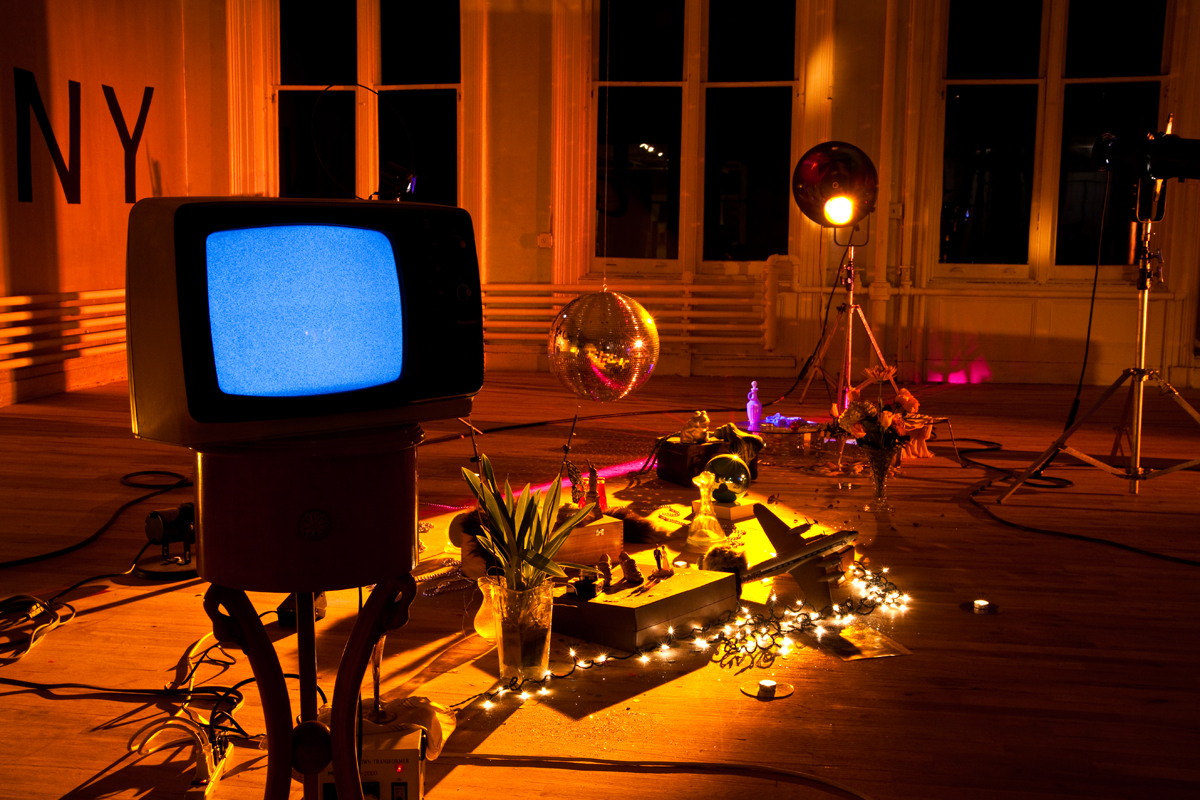 Objects are cluttered on the floor of a room lit with a warm, yellow hue. There is a low-hanging disco ball, two standing lights, and a small television showing white static mounted on a stand. There are two vases with plants and a string of white lights.