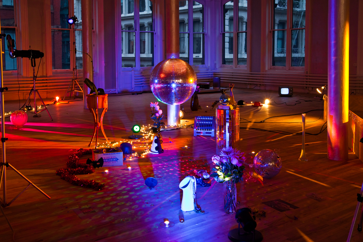 An installation shot of a room warmly lit by yellow and pink light. There is a low-hanging disco ball, surrounded by various objects set on the floor, including a vase with flowers, a second disco ball, strands of string lights, and more.